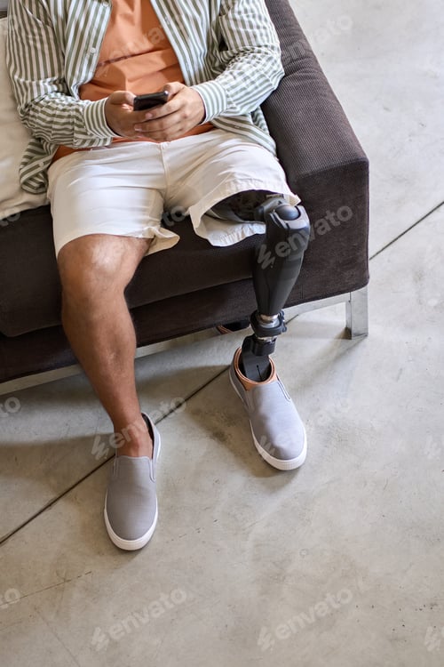 Preview: Amputee man with leg prosthesis sitting on sofa at home using phone.