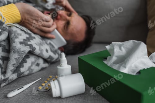 Preview: electronic thermometer and containers with treatment near sick man lying on couch and calling on