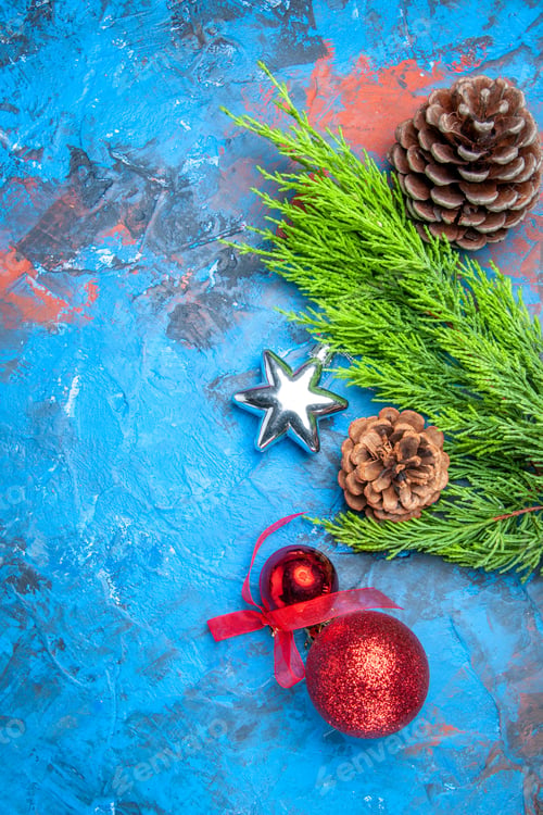 Preview: top view pine tree branch with pinecones and colorful xmas tree toys on blue-red background with