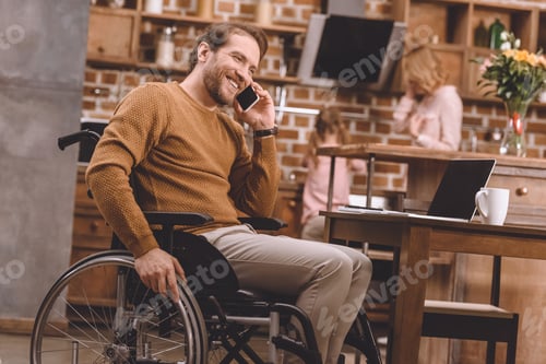 Preview: smiling disabled man in wheelchair talking on smartphone and using laptop with blank screen at home