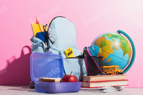 Preview: school backpack full of stationery near lunch box, globe, school bus model and books on pink