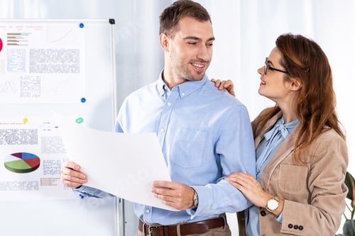 Preview: cheerful woman in glasses looking at handsome man with blank paper near white board with charts and