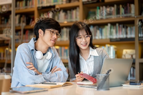Preview: Glasses asian woman student holding book and looking at laptop with a man friend at table in library