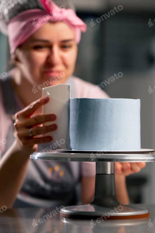 Preview: female baker in a professional kitchen distributes blue cream onto a sponge cake with a spatula