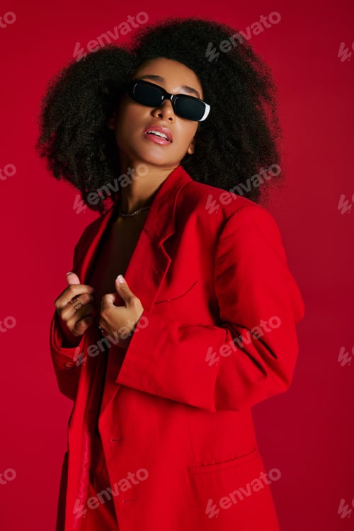 Preview: alluring african american woman in red unbuttoned blazer with sunglasses posing on red backdrop