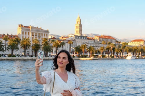 Preview: Portrait of beautiful young woman taking a selfie with beautiful coastal city of Split in background