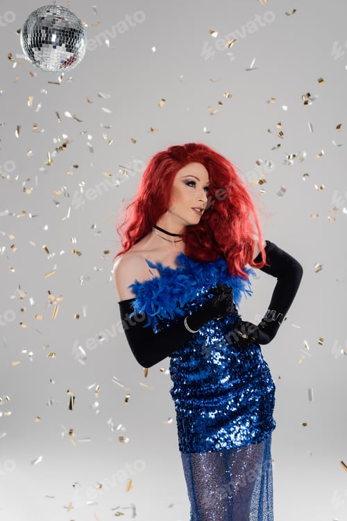 Preview: Glamorous drag queen in dress and gloves standing under confetti and disco ball on grey background