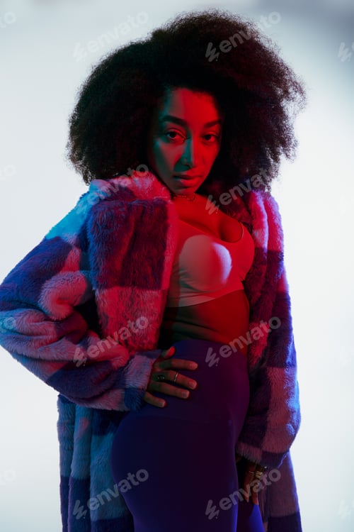 Preview: debonair african american woman in vibrant faux fur with curly hair looking at camera in lights