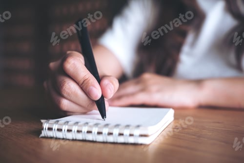 Preview: Closeup image of a woman's hand writing down on a white blank notebook on wooden table