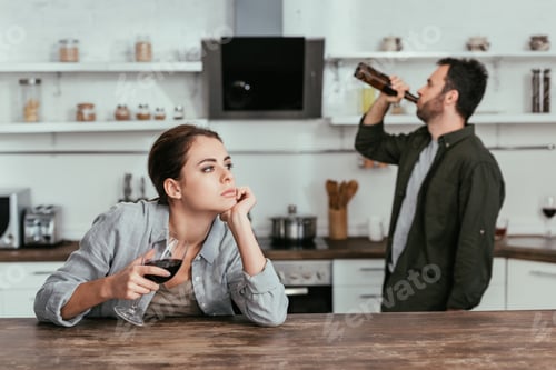Preview: Selective focus of pensive woman with wine glass and husband drinking beer at kitchen