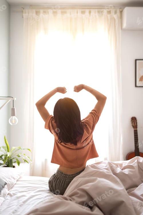 Preview: Rear view vertical portrait of unrecognizable woman waking up stretching arms sitting on bed ledge.