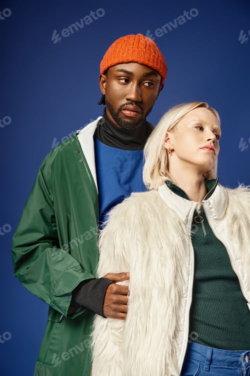 Preview: blonde woman posing with african american man in winter attire, diverse models on blue backdrop