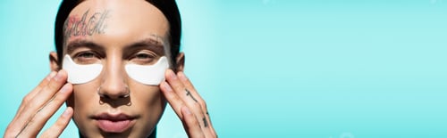 Preview: close up of tattooed young man adjusting eye patches and looking at camera isolated on turquoise,