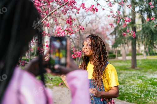Preview: African woman smiling while her friend takes a photo among pink blossoms.