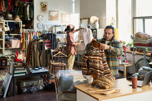 Preview: Young Adult Male Caucasian Shop Assistant Sorting Clothing in Thrift Store