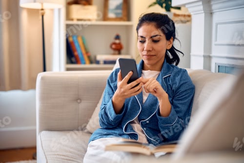 Preview: Latin American woman in pajama using cell phone while reading a book at home.