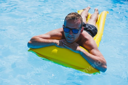 Preview: Attractive young man in sunglasses and shorts is relaxing on an inflatable yellow mattress