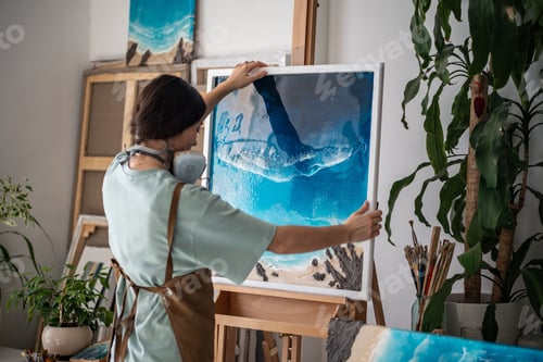 Preview: Thoughtful creative woman artist places finished epoxy painting on wooden easel in art workshop.