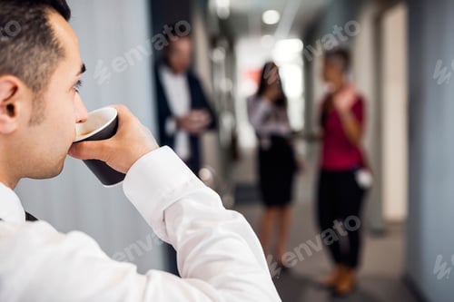 Preview: Businessman enjoying a coffee break while colleagues chat in the background.