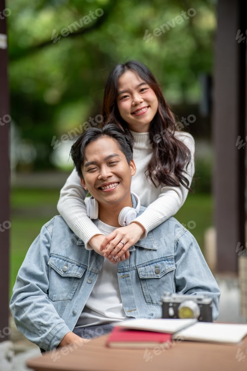 Preview: Young asian couple smiling woman standing and hugging man's shoulder while sitting at outdoor table