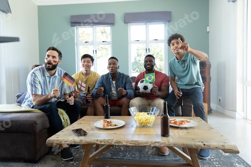 Preview: Curious multiracial male friends with flags, ball watching match while enjoying weekend at home