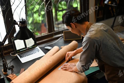 Preview: Handsome male artisan is meticulously measuring and cutting a large piece of leather on workbench