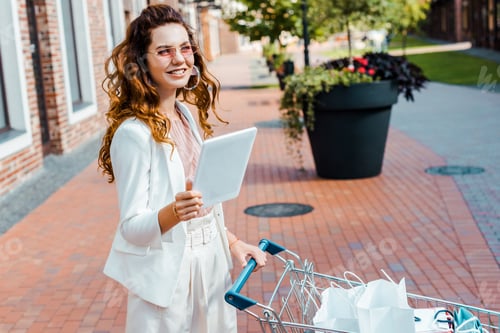 Preview: beautiful happy woman with shopping cart full of paper bags using tablet