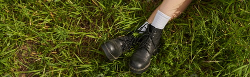 Preview: Top view of legs of woman in stylish boots sitting on grassy meadow outdoors, natural landscape