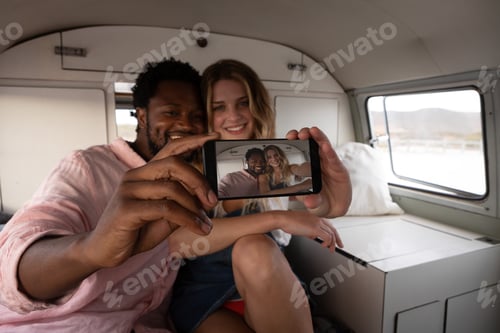 Preview: Front view of a multi-ethnic couple taking selfie in camper van at beach on a sunny day