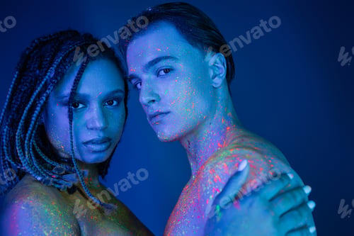 Preview: youthful multicultural couple with bare shoulders, in colorful neon body paint looking at camera