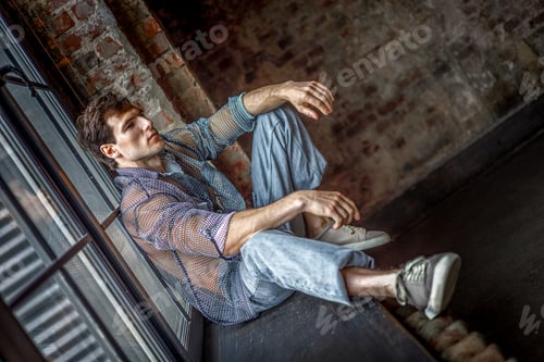 Preview: A modern young man with a stylish hairstyle sits on a windowsill near a window on a dark background.