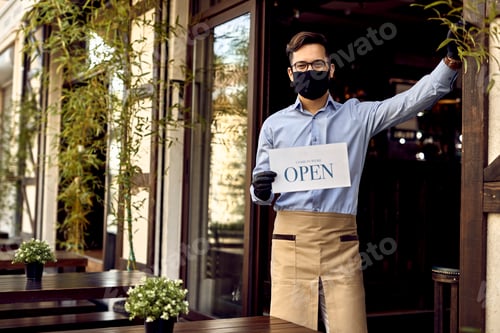 Preview: Happy waiter with protective face mask holding open sign while standing at doorway.