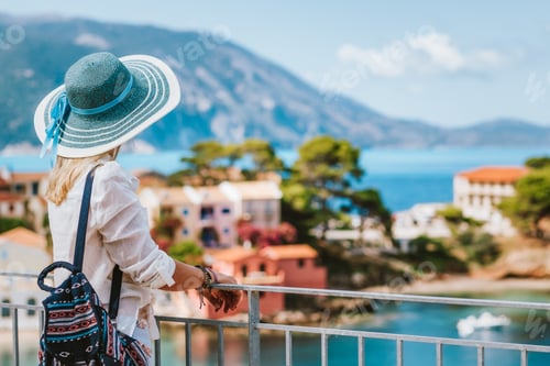 Preview: Tourist woman wearing blue sun hat enjoying top view of colorful tranquil village Assos on sunny day