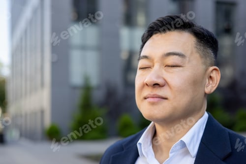 Preview: Close-up photo of a young Asian male office worker standing outside, thoughtfully closing his eyes