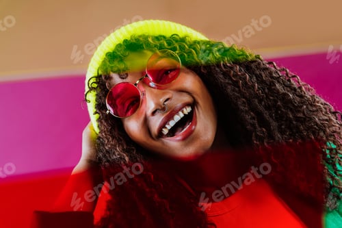 Preview: Young woman wearing sunglasses and beanie smiling with colorful background