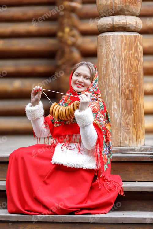 Preview: A girl in red traditional Russian clothes holds bagels in the spring near a wooden house