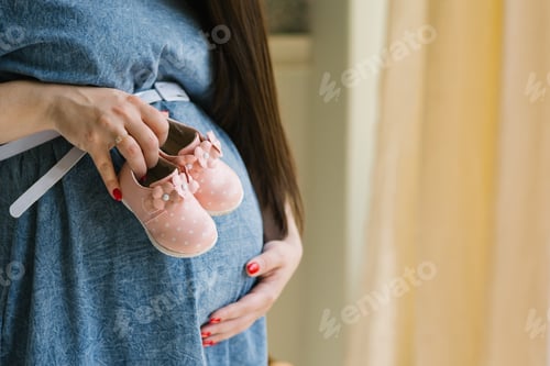 Preview: Closeup of Pregnant Woman Holding Pink Baby Booties on her Stomach in Denim Dress. Copy Space