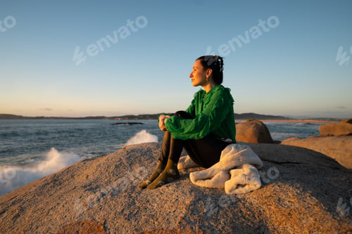 Preview: Young woman perched on seaside rock at punta faxilda, basking in golden sunset glow