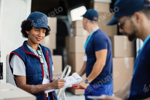 Preview: Smiling black delivery woman taking notes while talking to her coworker.