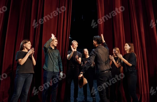 Preview: Standing against red curtains. Group of actors in dark colored clothes on rehearsal in the theater