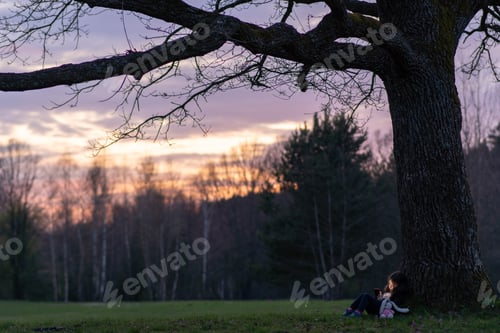 Preview: Child in huge world. Little girl with toy is sitting leaning large tree, looking at her phone