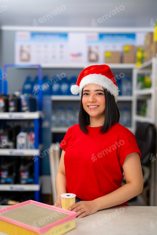Preview: Young woman smiling, wearing a santa hat while holding a car filter in an auto parts store