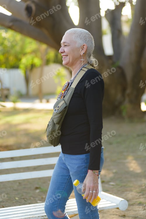 Preview: Middle-aged lesbian woman walks through a park carrying a bottle of juice in one hand.