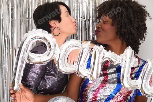 Preview: Two multiracial women in a party with a party silver balloon doing kissy face.