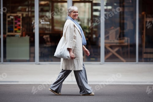 Preview: Elderly woman with short gray hair walks city street in fashionable attire pants, coat, sneakers