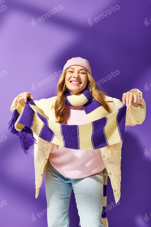 Preview: Joyful young woman in cozy winter outfit poses playfully against vibrant purple wall