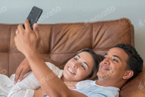 Preview: Happy young couple laying on the sofa at home and have fun using mobile phone