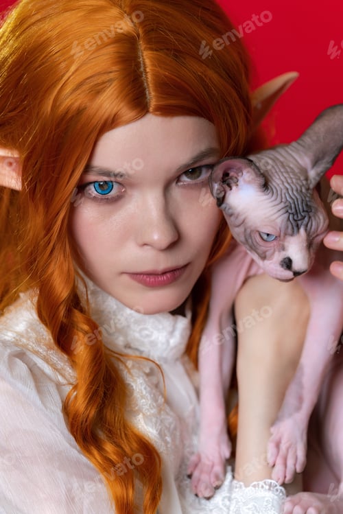 Preview: Portrait of young woman cosplay elf with red hair in white dress holding half asleep Sphinx kitten