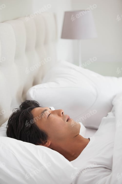 Preview: Asian man lying on bed in bedroom with upholstered headboard, white duvet and gray lamp