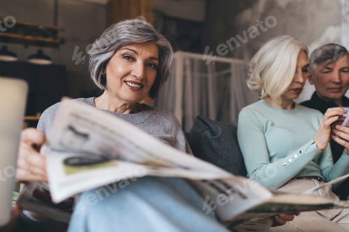 Preview: Cheerful senior woman reading newspaper while spending time with friends